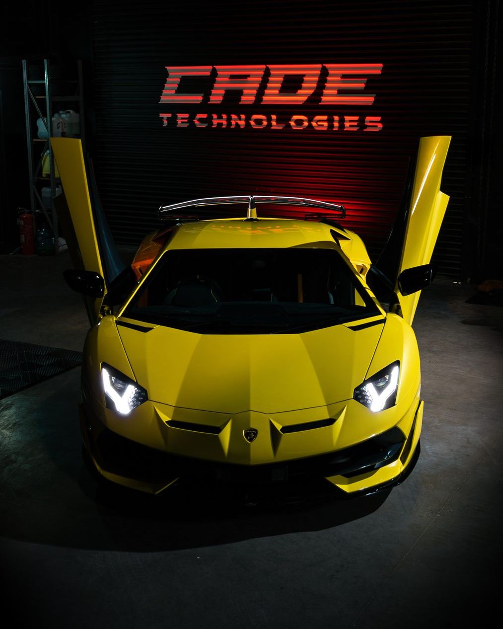 Yellow Lamborghini Aventador SVJ with its scissor doors raised beneath the illuminated CADE Technologies sign in the workshop
