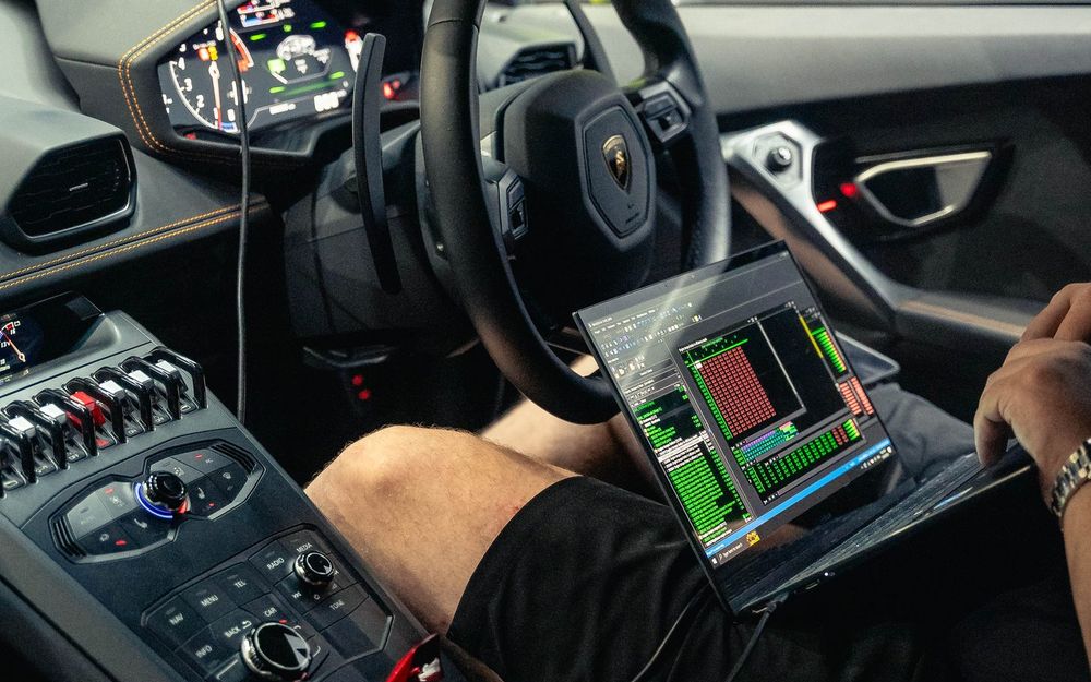 A tuner in the driver's seat of a Lamborghini Huracán with a laptop running flashing software, connected to the car by an OBD cable