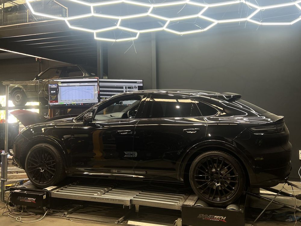 Black performance SUV strapped down on the Dynotech AWD dyno under hexagonal LED lights, with the dyno monitor beside it