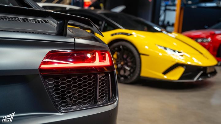 Taillight of a matte grey Audi R8 in the foreground with a yellow Lamborghini Huracán behind it in the workshop