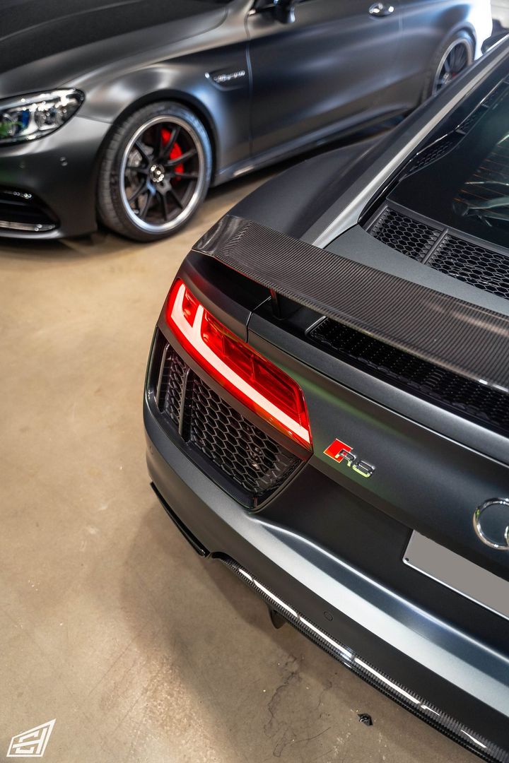 Rear of a matte grey Audi R8 with a carbon wing, beside a matte grey Mercedes-AMG coupe in the workshop