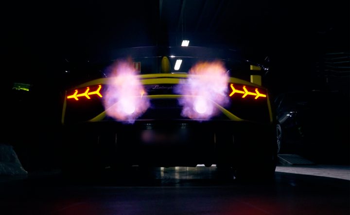 Rear view of a yellow Lamborghini Aventador SVJ in the dark, with flames from both exhaust outlets between its lit Y-shaped tail-lights