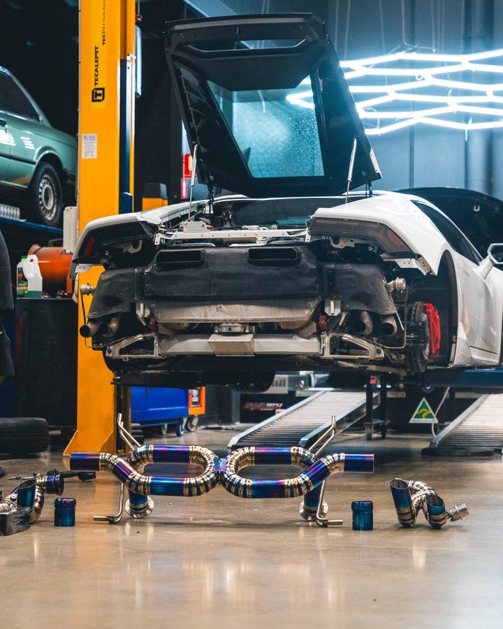 White Lamborghini Huracán raised on a hoist with its rear bumper removed and titanium exhaust parts laid out on the floor