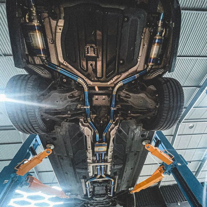 Full titanium exhaust system with heat-coloured blue and gold piping, seen from under the car on the CADE workshop hoist