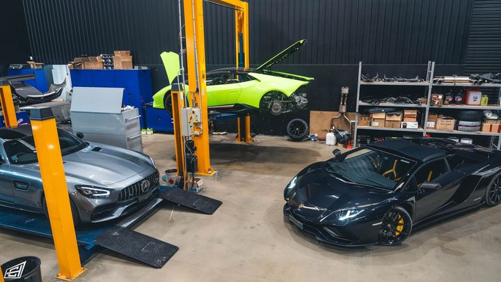 Workshop view with a grey Mercedes-AMG GT, a black Lamborghini Aventador and a green Lamborghini raised on a hoist