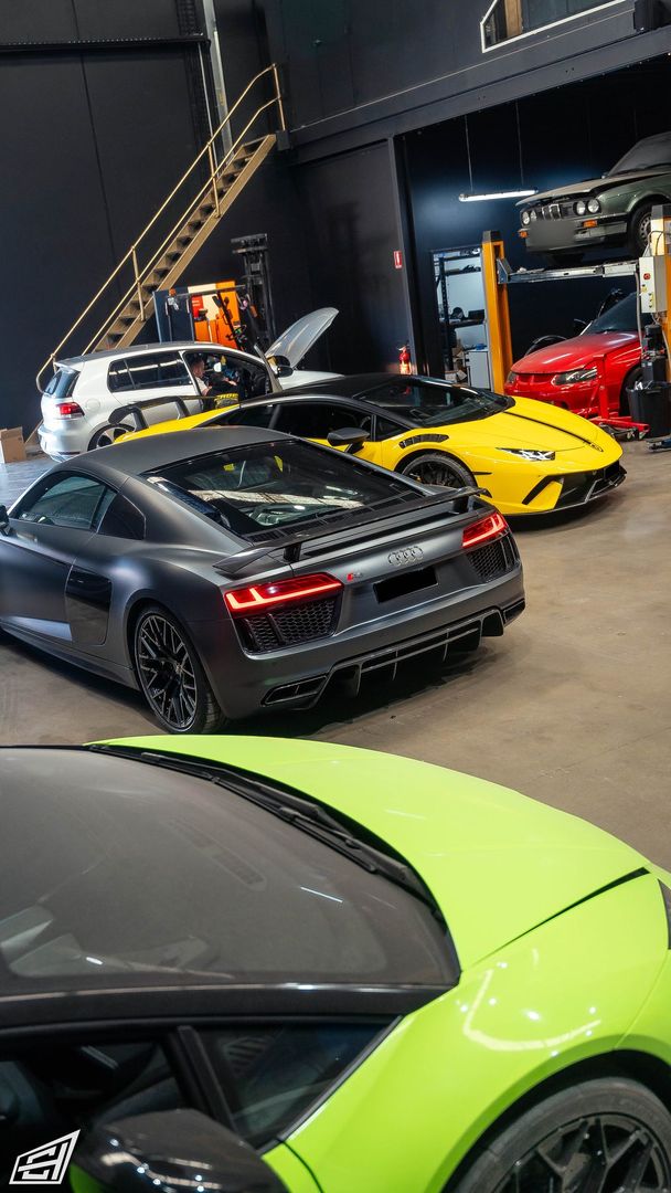 Workshop line-up with a matte grey Audi R8, a yellow Lamborghini and a red car, with a green Lamborghini in the foreground