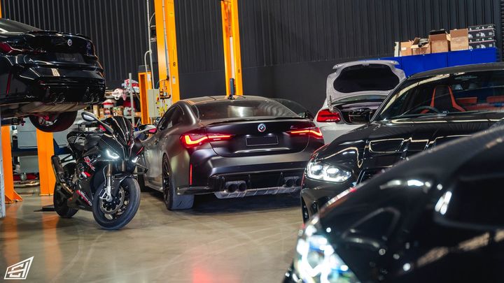 Workshop scene with a matte grey BMW coupe, a sport motorcycle and other cars, one raised on a hoist