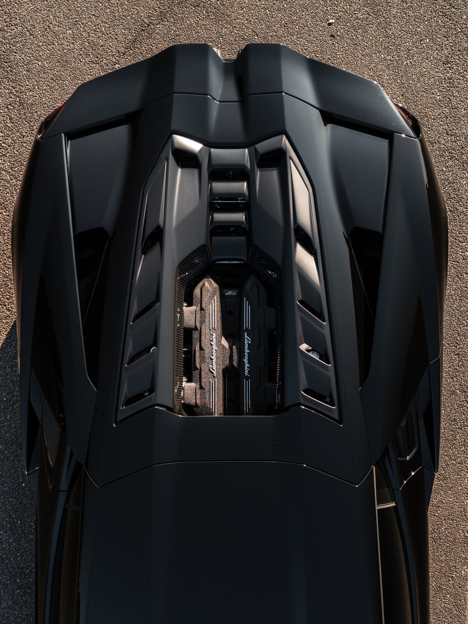 Overhead view of a black Lamborghini Revuelto's rear deck and exposed V12 engine covers on sunlit asphalt