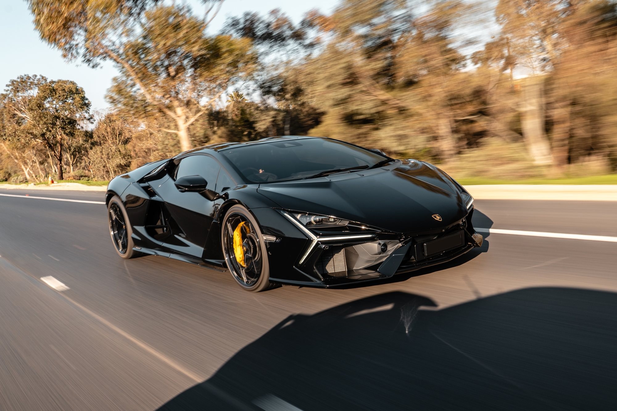 Black Lamborghini Revuelto with yellow brake calipers driving along a tree-lined road, front three-quarter rolling shot