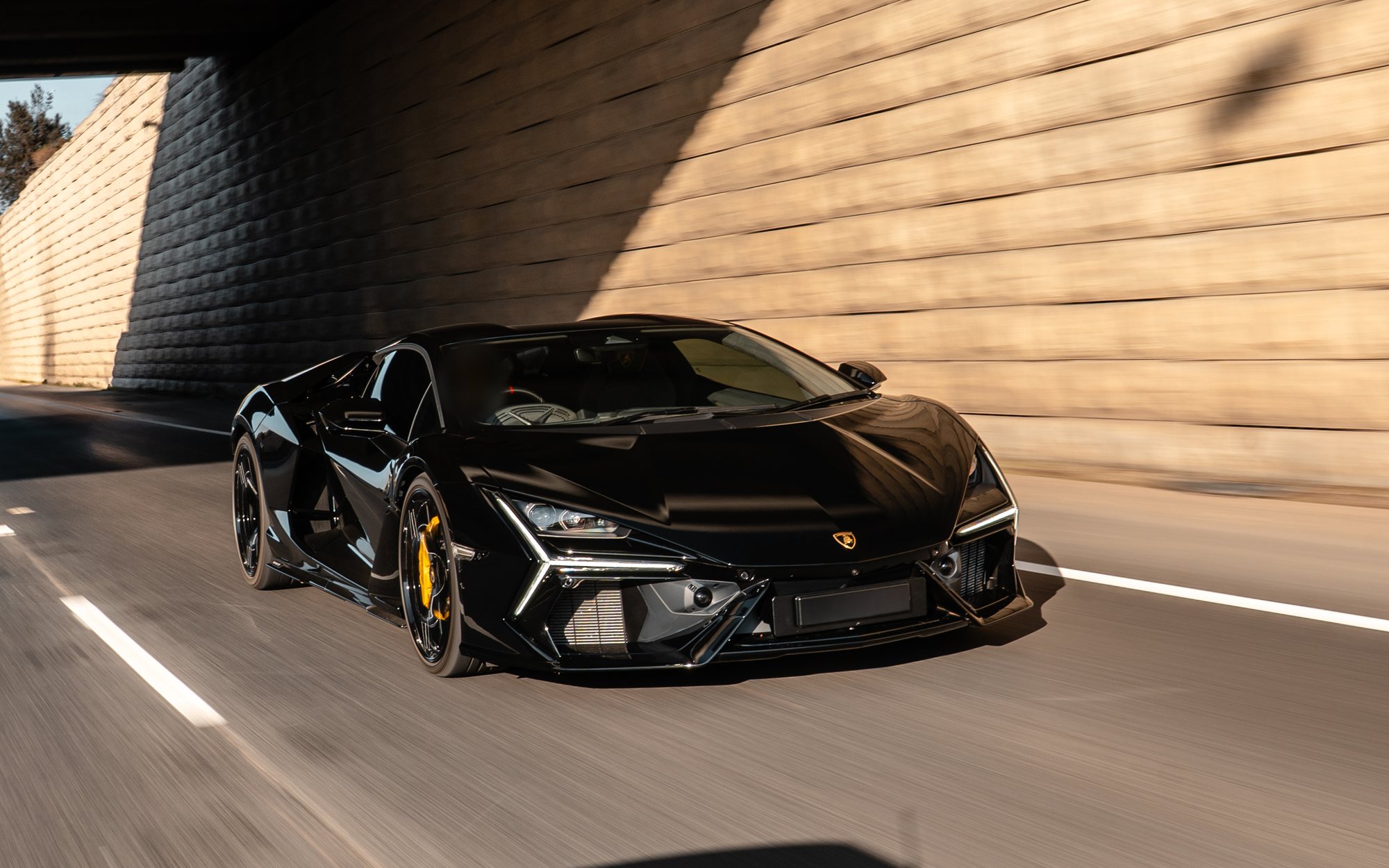 Black Lamborghini Revuelto with yellow brake calipers driving under an overpass beside a sunlit block wall, front three-quarter view