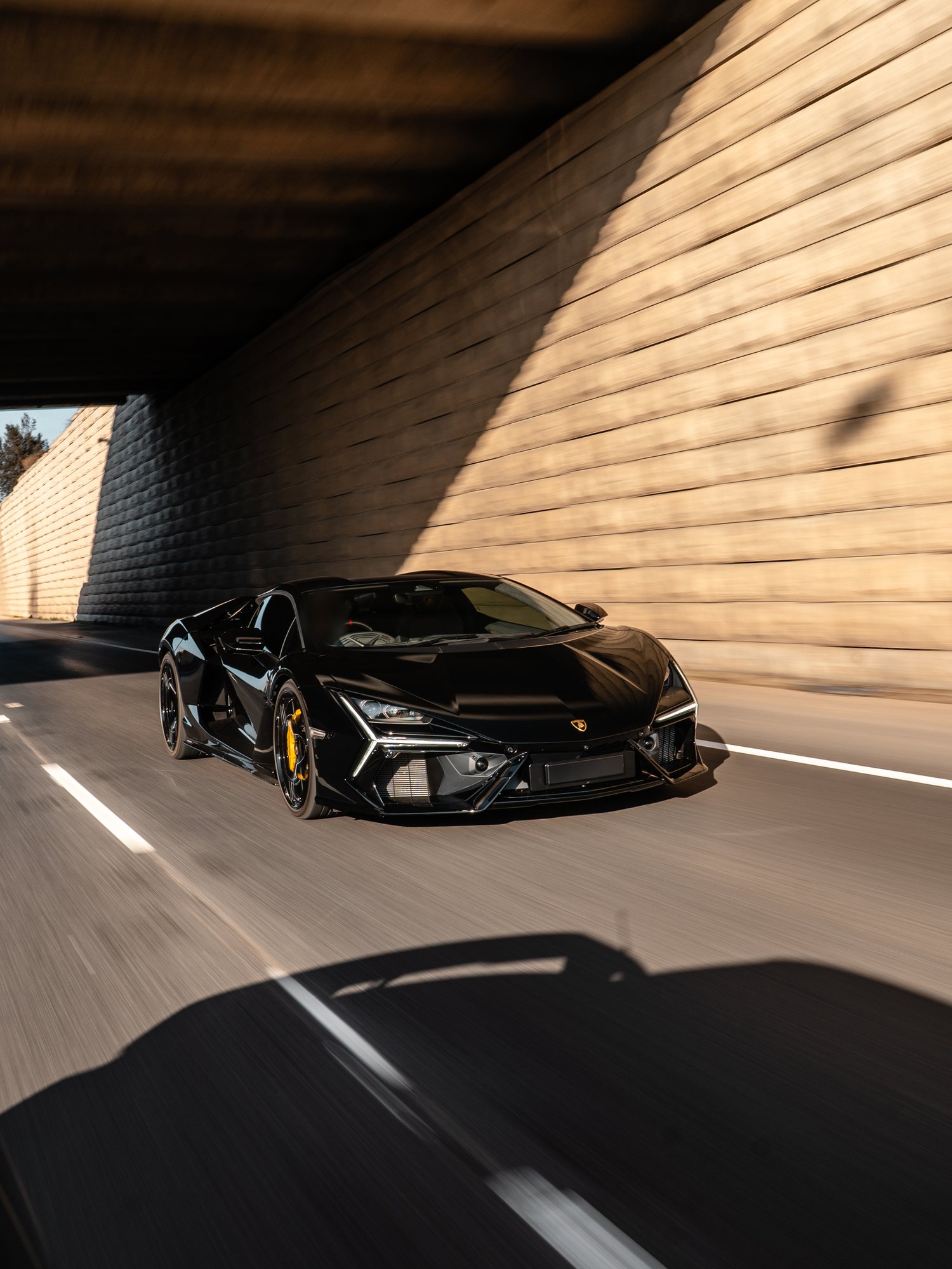 Black Lamborghini Revuelto with yellow brake calipers driving under a concrete overpass beside a sunlit block wall, front three-quarter rolling shot