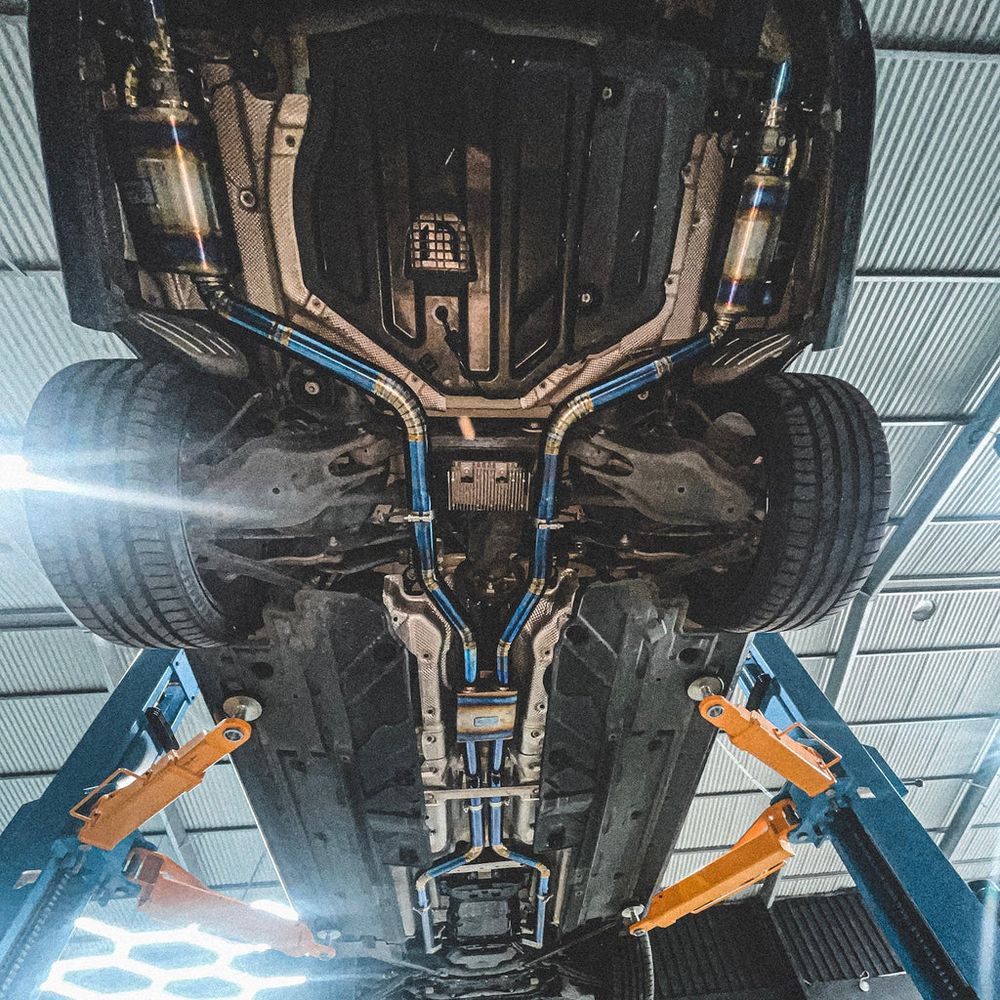 Full titanium exhaust system with heat-coloured blue and gold piping, seen from under the car on the CADE workshop hoist